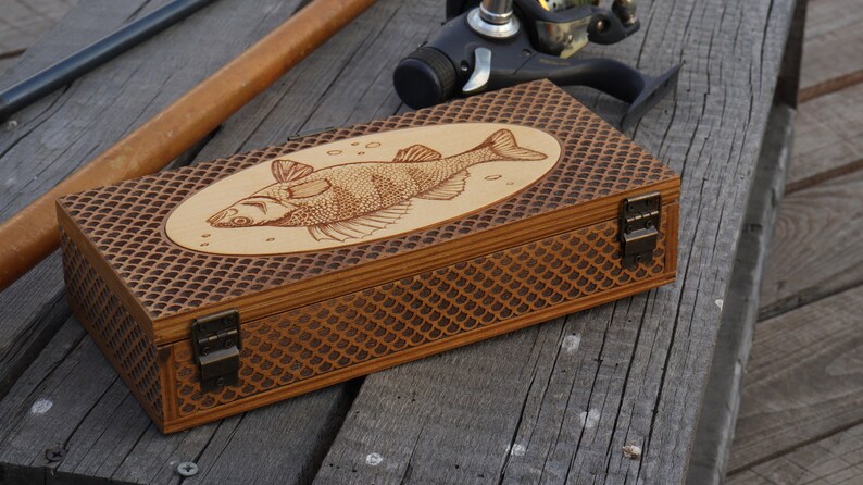 Op de afbeelding: Een houten doos met een visontwerp op de deksel. De doos heeft een bruine afwerking en een schubbenpatroon aan de zijkanten. De doos heeft twee messing sluitingen.