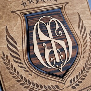 Puede incluir: Una placa de madera con un dise&ntilde;o en forma de escudo que presenta un monograma de las letras "S" y "D" en una fuente decorativa. El escudo est&aacute; rodeado por una corona de laurel y tres estrellas.