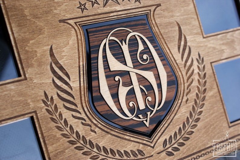 Puede incluir: Una placa de madera con un dise&ntilde;o en forma de escudo que presenta un monograma de las letras "S" y "D" en una fuente decorativa. El escudo est&aacute; rodeado por una corona de laurel y tres estrellas.