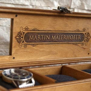 May include: A wooden watch box with a glass top and a personalized inscription that reads "MARTIN MAIERHOFER". The box has a velvet interior with compartments for storing watches.