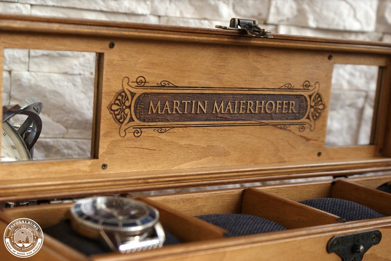 May include: A wooden watch box with a glass top and a personalized inscription that reads "MARTIN MAIERHOFER". The box has a velvet interior with compartments for storing watches.