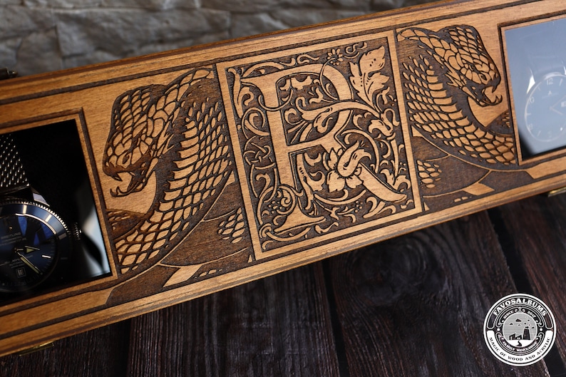 May include: A wooden watch box with a dark brown stain. The box has three compartments, each with a glass window. The compartments are decorated with laser-engraved designs of snakes and a letter "R" with floral accents. The box is made by "Favos Albums, Made of Wood and Acrylic."