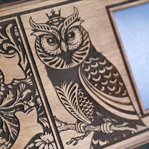 May include: A wooden picture frame with a decorative design of a crowned owl and floral patterns. The frame has a blank space for a photo.