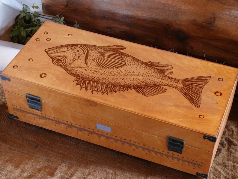 May include: A wooden box with a fish design engraved on the lid. The box has a ruler on the side and a metal latch.
