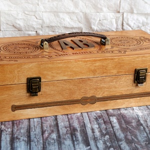 May include: Wooden box with a brown leather handle and metal hardware. The box is engraved with decorative elements and the initials "AB". The text "I'M A HOOKER ON ONE MORE CAST" is also visible.