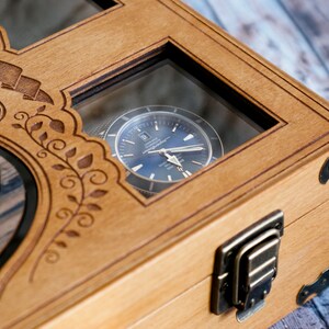 May include: A wooden watch box with a glass window showing a silver watch with a blue face. The box has a decorative design with a shield and floral patterns. The box has two brass clasps.