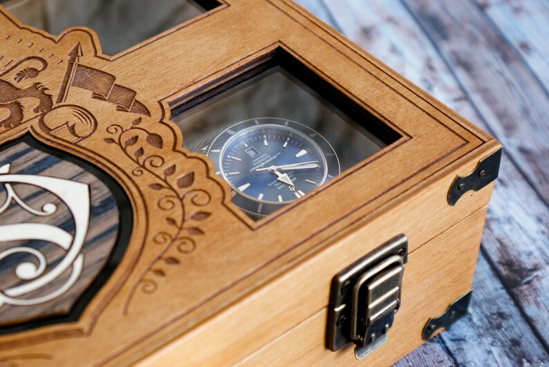 May include: A wooden watch box with a glass window showing a silver watch with a blue face. The box has a decorative design with a shield and floral patterns. The box has two brass clasps.
