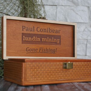 Op de afbeelding: Een houten doos met een scharnierend deksel. Het deksel is open en heeft een gepersonaliseerde inscriptie met de tekst "Paul Conibear, Lundin Mining, Gone Fishing!" De doos heeft een geweven patroon op de bodem.