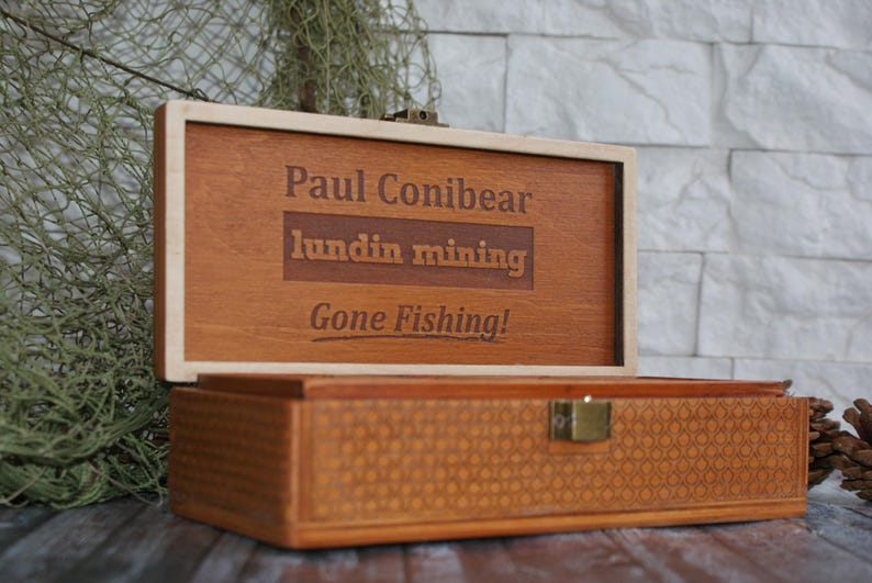 Op de afbeelding: Een houten doos met een scharnierend deksel. Het deksel is open en heeft een gepersonaliseerde inscriptie met de tekst "Paul Conibear, Lundin Mining, Gone Fishing!" De doos heeft een geweven patroon op de bodem.