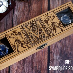 May include: A wooden watch box with a clear glass lid. The box is decorated with an engraved bull and the letter "M". The box is labeled "Gift with the Symbol of 2021 - Bull".