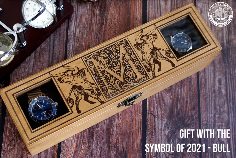 May include: A wooden watch box with a clear glass lid. The box is decorated with an engraved bull and the letter "M". The box is labeled "Gift with the Symbol of 2021 - Bull".