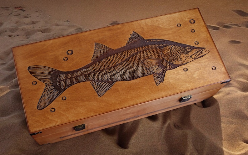 K&ouml;nnte beinhalten: Eine Holzkiste mit einem Scharnierdeckel, auf dem ein Laser-graviertes Bild eines Fisches mit Blasen zu sehen ist. Die Kiste hat zwei Messing-Scharniere und einen Messing-Verschluss.