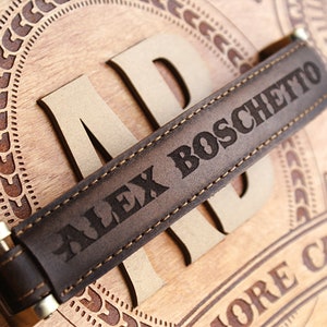 May include: Close-up of a brown leather strap with the name "ALEX BOSCHETTO" engraved in capital letters. The strap is stitched with light-colored thread and has gold-colored metal accents. The strap is placed on a wooden surface with carved details.