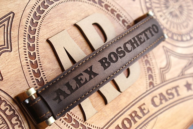 May include: Close-up of a brown leather strap with the name "ALEX BOSCHETTO" engraved in capital letters. The strap is stitched with light-colored thread and has gold-colored metal accents. The strap is placed on a wooden surface with carved details.