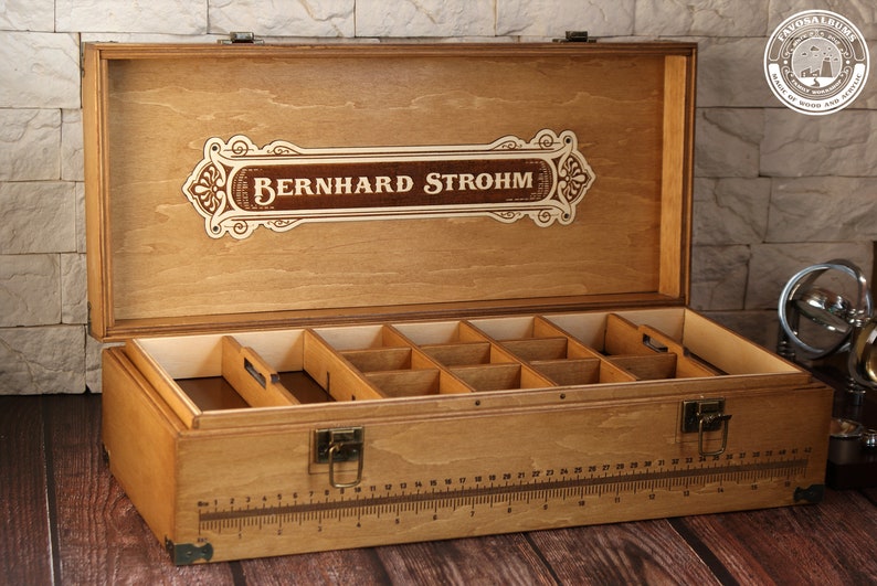 May include: A wooden box with a hinged lid and a label that reads "Bernhard Strohm". The box has a ruler on the front and is divided into compartments inside.