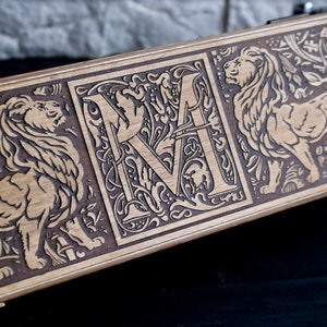 May include: A wooden box with a decorative design featuring a lion, a letter "M" surrounded by floral motifs, and another lion. The box is made of light brown wood and has a dark brown stain.