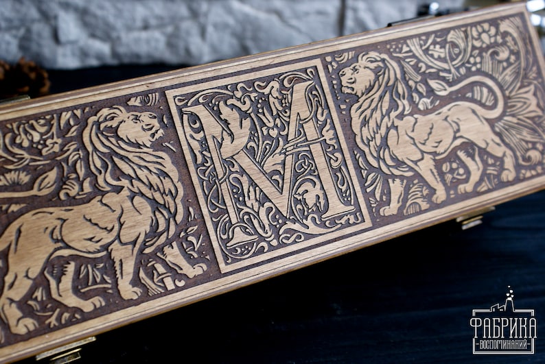 May include: A wooden box with a decorative design featuring a lion, a letter "M" surrounded by floral motifs, and another lion. The box is made of light brown wood and has a dark brown stain.