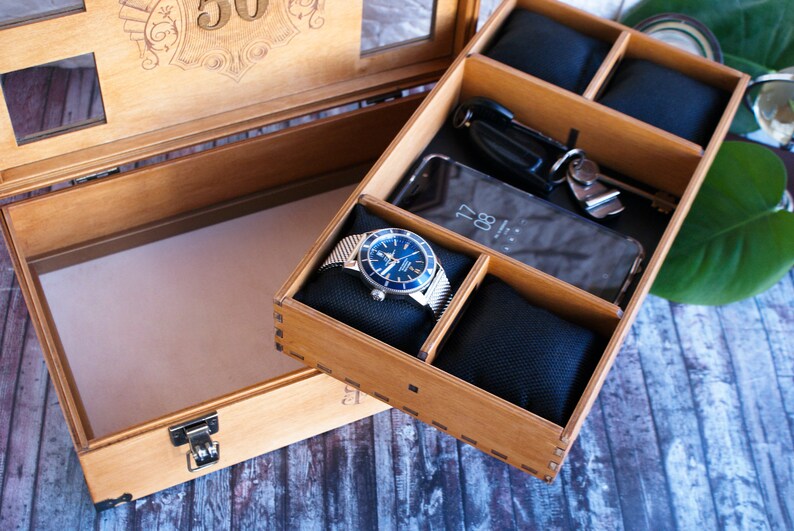 Puede incluir: Una caja de madera con una tapa que se abre para revelar un compartimento con un reloj, un tel&eacute;fono y un llavero. La caja est&aacute; hecha de madera clara y tiene un acabado natural. La caja est&aacute; decorada con el n&uacute;mero "30" en la tapa.