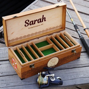 May include: A rectangular wooden fishing tackle box with a hinged lid, featuring the name Sarah and Bridgehampton, NY printed inside. The interior has green felt-lined compartments, and the exterior displays wave carvings and a life ring emblem.