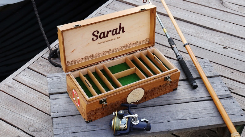 May include: A rectangular wooden fishing tackle box with a hinged lid, featuring the name Sarah and Bridgehampton, NY printed inside. The interior has green felt-lined compartments, and the exterior displays wave carvings and a life ring emblem.