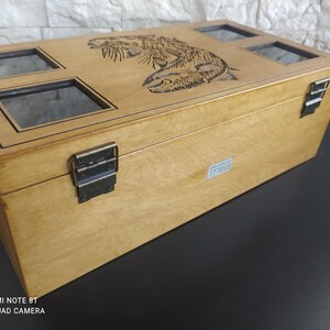 May include: A light brown wooden box with a carved fish design on the lid. The box features four glass windows and metal clasps. The rectangular box is likely a storage container or display case.
