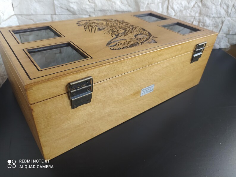May include: A light brown wooden box with a carved fish design on the lid. The box features four glass windows and metal clasps. The rectangular box is likely a storage container or display case.