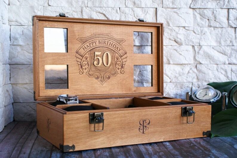 Puede incluir: Una caja de madera con tapa abatible y un interior de terciopelo. La tapa est&aacute; grabada con las palabras "Happy Birthday 50". La caja tiene un cierre y bisagras de lat&oacute;n.