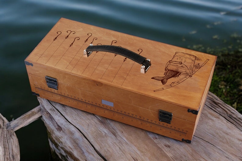 May include: A wooden fishing tackle box with a leather handle. The box has a ruler on the side and a fish and bucket design on the lid.