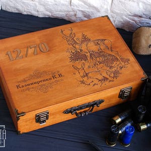 May include: A wooden box with a hinged lid, engraved with the number "12/70", the name "Kazimireko K.B.", and a design of deer in a forest. The box has a brass handle and metal clasps.