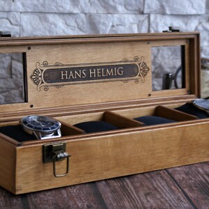 May include: A wooden watch box with a glass top and a personalized inscription that reads "Hans Helmlig". The box has four compartments lined with black velvet and a brass latch.