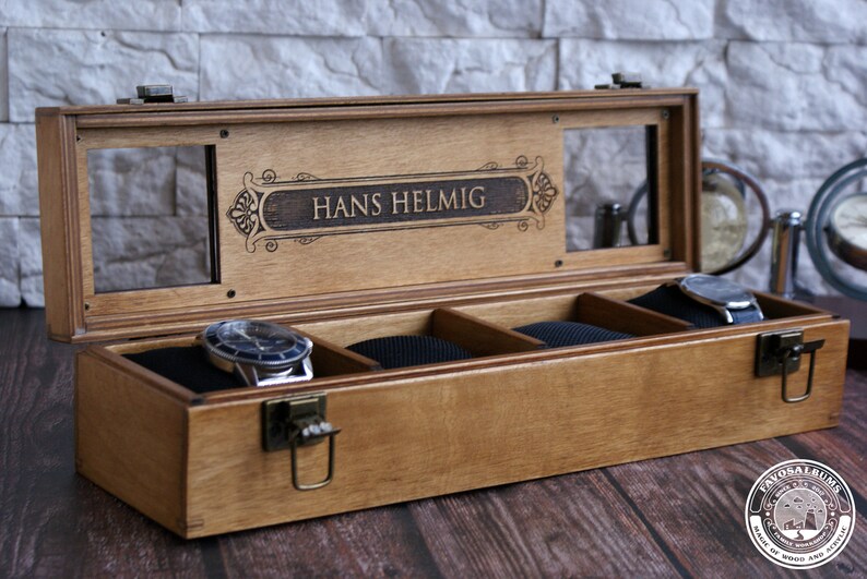 May include: A wooden watch box with a glass top and a personalized inscription that reads "Hans Helmlig". The box has four compartments lined with black velvet and a brass latch.