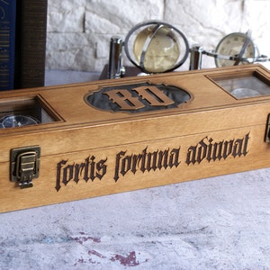 Peut inclure: Une bo&icirc;te rectangulaire en bois avec deux fen&ecirc;tres en verre, des loquets en m&eacute;tal couleur laiton et le texte latin grav&eacute; "fortis fortuna adiuvat" sur le devant. Le couvercle pr&eacute;sente une plaque d&eacute;corative centrale avec les lettres "BD".