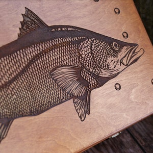 K&ouml;nnte beinhalten: Eine Holzkiste mit einem lasergravierten Bild eines Fisches. Der Fisch ist detailliert mit Schuppen und Flossen dargestellt. Die Kiste ist hellbraun und hat eine glatte Oberfl&auml;che.