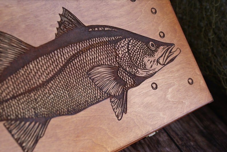 K&ouml;nnte beinhalten: Eine Holzkiste mit einem lasergravierten Bild eines Fisches. Der Fisch ist detailliert mit Schuppen und Flossen dargestellt. Die Kiste ist hellbraun und hat eine glatte Oberfl&auml;che.