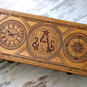 May include: A wooden box with a decorative design featuring a clock, compass, and monogrammed initials. The box is made of light wood and has a natural finish.