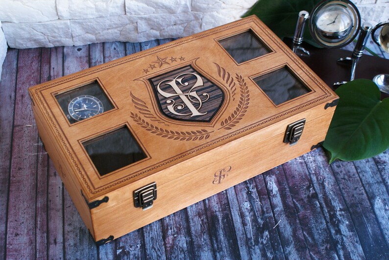 Puede incluir: Una caja de madera para relojes con tapa de cristal transparente y un dise&ntilde;o grabado personalizado. La caja tiene un cierre de lat&oacute;n y est&aacute; decorada con un escudo y una corona de laurel.
