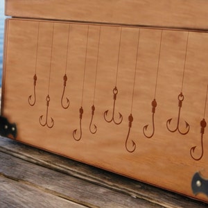 May include: A wooden box with a brown stain and metal corner accents. The lid of the box is decorated with an engraved design of multiple fishing hooks.