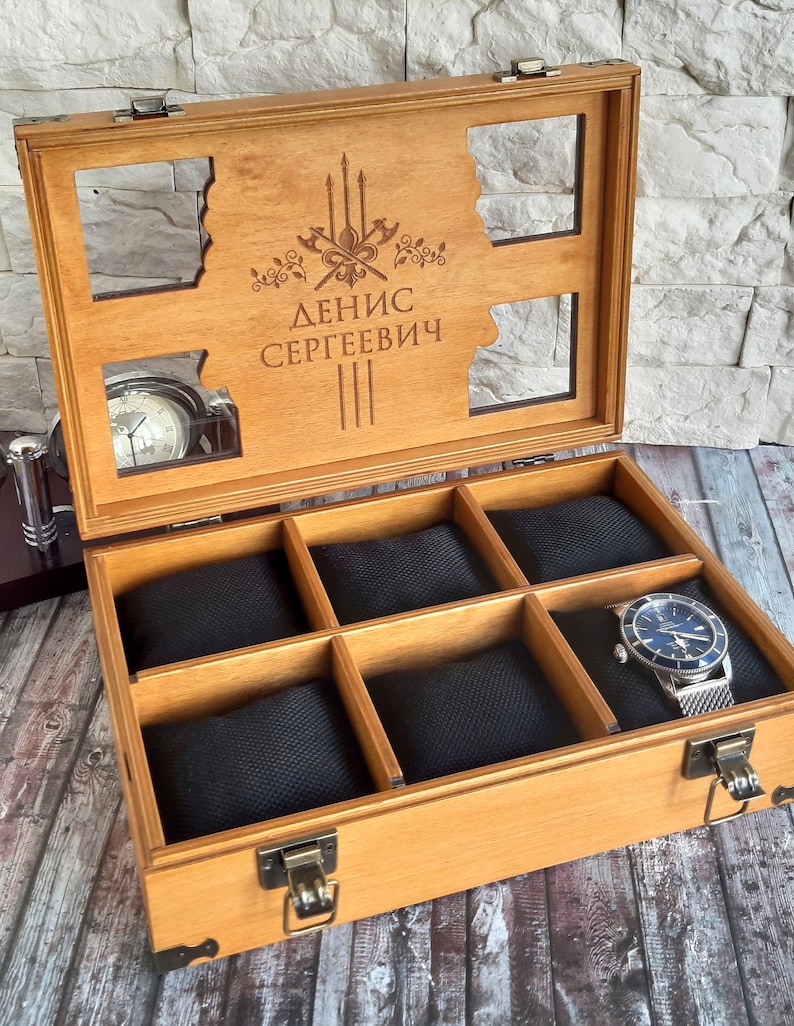 May include: A wooden watch box with a clear lid and six compartments lined with black fabric. The lid has an engraved design with the text "ДЕНИС СЕРГЕЕВИЧ".