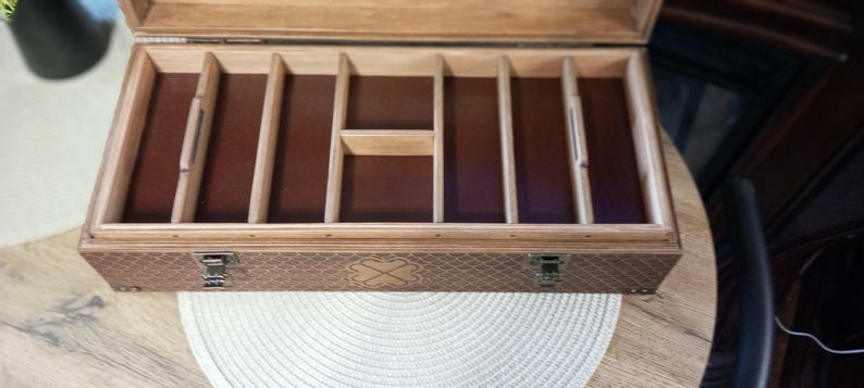 May include: Open wooden box with multiple compartments and a hinged lid. The interior features a dark brown lining and dividers. The exterior has a textured pattern and a metal clasp closure. The box is designed for storage.