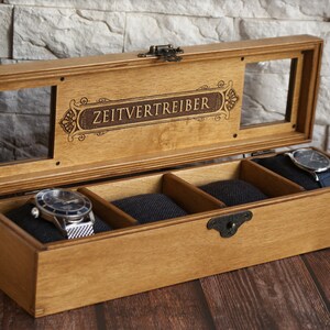 May include: A wooden watch box with a glass top and four compartments. The box is engraved with the words "Zeitvertreiber".