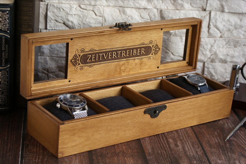 May include: A wooden watch box with a glass top and four compartments. The box is engraved with the words "Zeitvertreiber".