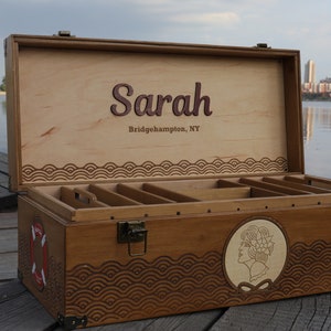 May include: A rectangular wooden storage chest with a hinged lid, featuring the name Sarah and Bridgehampton, NY engraved inside. The exterior has a dark wood finish, metal latches, a life preserver graphic, and a circular portrait engraving.