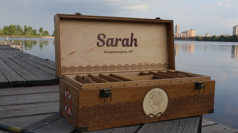 May include: A rectangular wooden storage chest with a hinged lid, featuring the name Sarah and Bridgehampton, NY engraved inside. The exterior has a dark wood finish, metal latches, a life preserver graphic, and a circular portrait engraving.