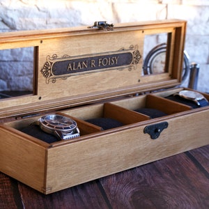 May include: A wooden watch box with a glass top and three compartments. The box is engraved with the name "ALAN R FOISY".