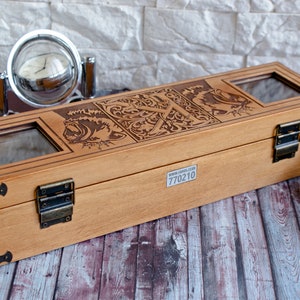 May include: A wooden watch box with a hinged lid and brass clasps. The box has a decorative engraved design on the top and a clear glass window on the lid. The box is sitting on a wooden surface. The text "770210" is printed on the bottom of the box.