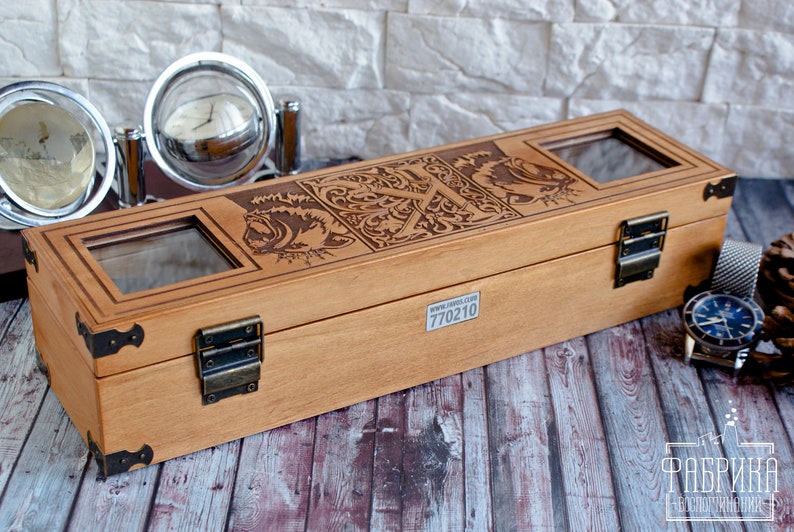 May include: A wooden watch box with a hinged lid and brass clasps. The box has a decorative engraved design on the top and a clear glass window on the lid. The box is sitting on a wooden surface. The text "770210" is printed on the bottom of the box.