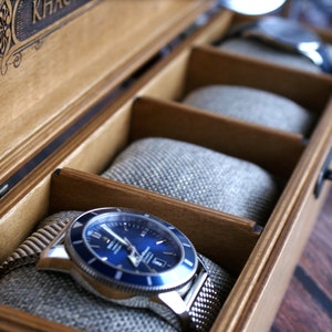 Pu&ograve; includere: Una scatola porta orologi in legno con un coperchio in vetro e un orologio blu con un cinturino argentato all'interno. La scatola ha un'etichetta che dice "KHAC"