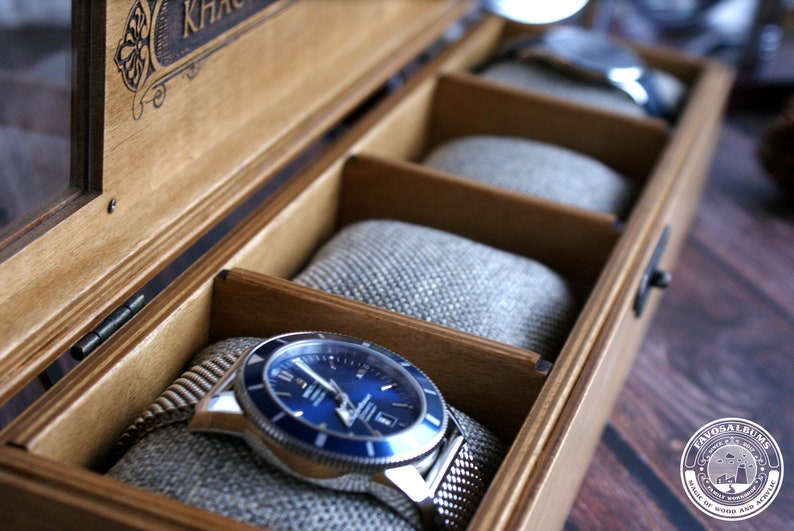 Pu&ograve; includere: Una scatola porta orologi in legno con un coperchio in vetro e un orologio blu con un cinturino argentato all'interno. La scatola ha un'etichetta che dice "KHAC"