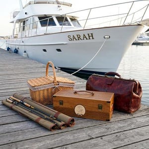 May include: A woven wicker picnic basket with a checkered liner, a rectangular wooden tackle box with a circular emblem, a dark brown leather doctor-style bag, and a rolled olive green canvas fishing rod case on a wooden dock near a white yacht.