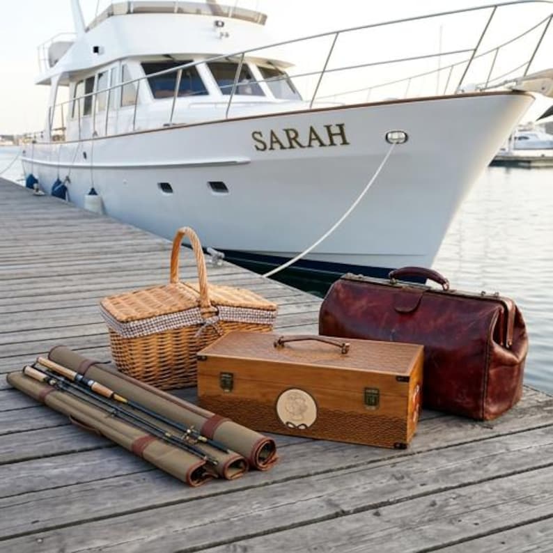 May include: A woven wicker picnic basket with a checkered liner, a rectangular wooden tackle box with a circular emblem, a dark brown leather doctor-style bag, and a rolled olive green canvas fishing rod case on a wooden dock near a white yacht.